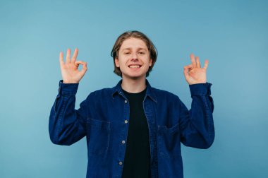 Tişörtlü olumlu bir adam mavi bir arkaplanda yüzünde bir gülümsemeyle duruyor ve parmaklarını gösteriyor.