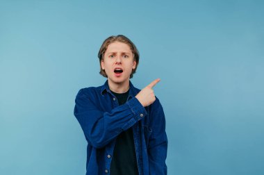 Şok olmuş bir yüz ifadesiyle mavi arka planda duran tişörtlü, şık bir adam kameraya bakıyor ve parmağını fotokopi uzayına doğrultuyor.