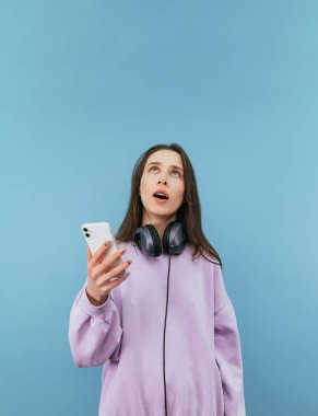 Ellerinde akıllı telefonlar, boynunda kulaklıklar olan sürpriz bir kadın mavi arka planda ağzı açık fotokopi odasına bakıyor. Dikey