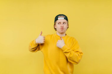 Eşofmanlı ve şapkalı olumlu bir adam sarı arka planda izole edilmiş komik bir yüzle başparmağını kaldırıyor ve başka tarafa bakıyor.