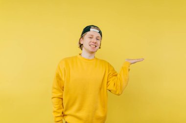 Sarı tişörtlü ve şapkalı mutlu adam sarı arka planda izole edilmiş elinde boş bir yer tutuyor ve yüzünde bir gülümsemeyle kameraya bakıyor..