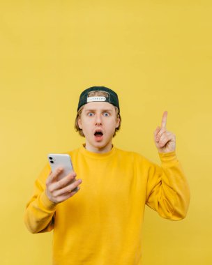Elinde akıllı telefon olan, sarı arka planda izole edilmiş, boş alanda baş parmağını gösteren ve kameraya bakan şok olmuş genç bir adamın portresi. Dikey.