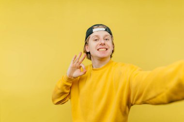 Sarı kazaklı ve şapkalı neşeli bir adam yüzünde bir gülümsemeyle selfie çeker ve sarı bir arka planda izole edilmiş, hoş bir jest gösterir..