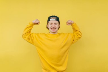 Parlak eşofmanlı neşeli adam bisepslerini gösteriyor ve yüzünde bir gülümsemeyle kameraya bakıyor, sarı bir arka planda izole edilmiş.