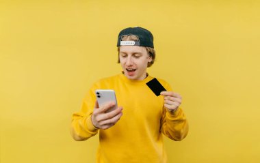 Sarı kazaklı ve şapkalı duygusal adam sarı arka planda izole edilmiş, elinde bir harita, telefon ekranına bakıyor..