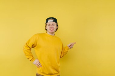 Yüzünde bir tebessümle sarı bir arka planda duran renkli günlük kıyafetli olumlu bir adam kameraya bakar ve parmağını yana çevirir.