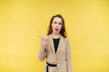 Şok olmuş ceketli kadın sarı arka planda duruyor ve parmağını yana doğrultuyor ve kameraya şaşırmış bir şekilde bakıyor..