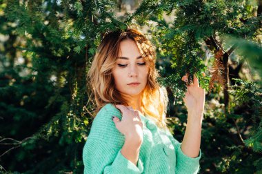 Süveter giyen çekici sarışın kameraya daldaki kozalaklı ağaçların arka planında poz veriyor.