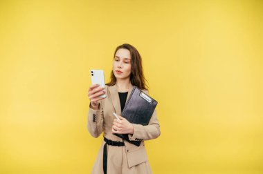 Ceketli, tabletli çekici bir iş kadını sarı arka planda duruyor ve ciddi bir yüzle akıllı telefon kullanıyor..