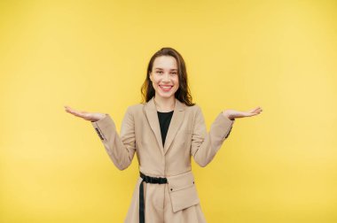 Ceketli, yüzünde bir gülümsemeyle pozitif bir kadın kollarını açar ve sarı arka planda izole bir şekilde kameraya bakar..