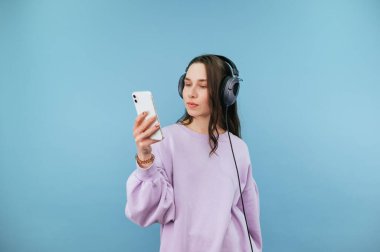 Kulaklıklı ve mor kazaklı çekici bir kadın mavi arka planda duruyor ve ciddi yüzlü bir akıllı telefon kullanıyor..