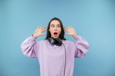 Şok olmuş yüzü, günlük kıyafetleri ve kulaklıkları boynuna dolanmış kolları havada, mavi arka planda soyutlanmış bir kadın.