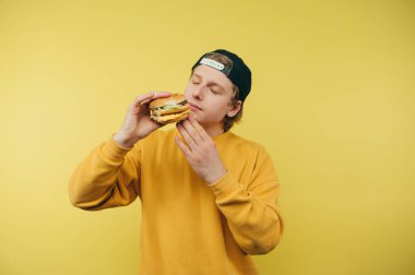 Şapkalı genç adam elinde kocaman lezzetli bir burgeri kokluyor sarı bir arka planda, fast food.