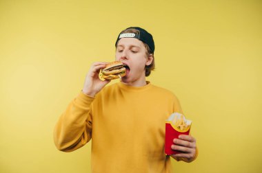 Sarı kazaklı genç bir adam sarı arka planda elinde patates kızartmasıyla duruyor ve iştah açıcı bir burger yiyor..