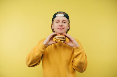 Sarı arka planda fast food yiyen yakışıklı bir adam. İştah açıcı bir hamburgerle izole edilmiş. Kameraya bakıyor ve gülümsüyor..