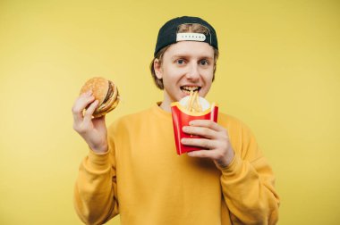 Elinde hamburgerle içeri giren komik adam sarı arka planda yüzünde bir gülümsemeyle patates kızartması yiyor. Genç adam ve fast food..
