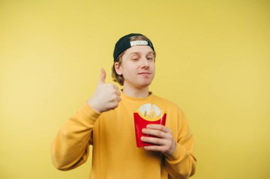 Pozitif genç adam elinde patates kızartması paketiyle sarı arka planda duruyor ve kameraya bakıyor ve ciddi bir yüzle baş parmağını kaldırıyor.