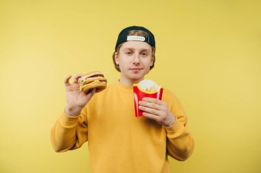 Elinde fast food tutan yakışıklı genç bir adam ve yüzünde bir gülümsemeyle kameraya bakıyor. Adam patates kızartması ve hamburger yiyor.