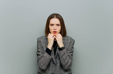 Şok olmuş yüzlü, ceketli ürkek iş kadını gri duvarın arka planındaki kameraya bakıyor..