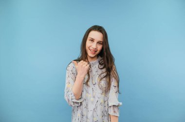 Dişlerinde diş teli olan mutlu bir kız gülümser ve mavi arka plandaki kameraya bakar, güzel bir elbise giyer ve parmağını yana çevirir..