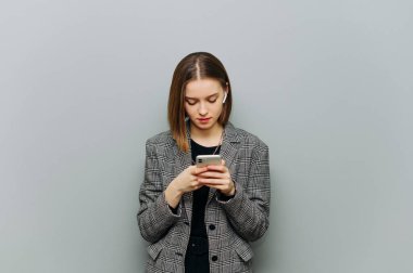 Ceketli konsantre bir kadın gri duvarda ciddi bir yüzü olan bir akıllı telefon kullanıyor..