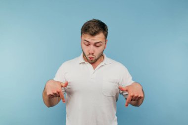 Beyaz tişörtlü, tüylü, şok olmuş bir yüz ve mavi arka planda izole edilmiş parmaklarını gösteren şaşırmış bir adam..