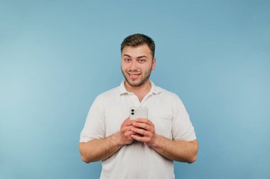 Beyaz tişörtlü, beyaz arka planda duran mavi arka planda elinde akıllı bir telefonla kameraya gülümseyen bir adam..