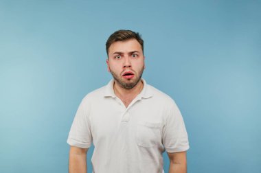 Mavi arka planda beyaz tişörtlü, şok olmuş ve korkmuş bir yüzle kameraya bakan şaşırmış bir adamın portresi.