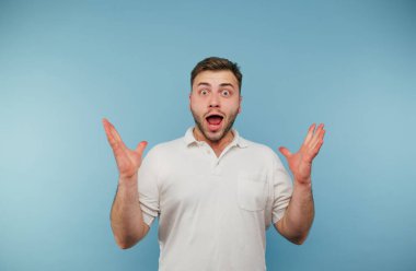 Beyaz tişörtlü, elleri havada ve mavi arka planda duygusal yüzlü şok olmuş bir adamın portresi..