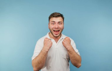 Neşeli yetişkin bir adamın portresi zaferle coşuyor. Eller havada kameraya bakıyor ve mavi arka planda izole edilmiş mutlulukla bağırıyor.