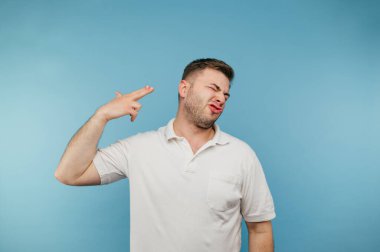 Beyaz tişörtlü adam kendini parmağıyla vurdu ve yüzünü buruşturdu. Yetişkin bir adam mavi arka plandaki bir silahın parmağından vurularak intihar etti..