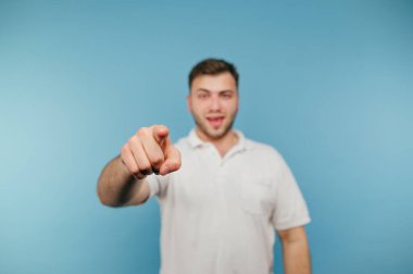 Beyaz tişörtlü pozitif adam kamerayı işaret ediyor ve gülümsüyor, mavi arka planda soyutlanmış.