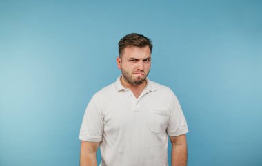 Beyaz tişörtlü tatminsiz bir adam mavi arka planda duruyor ve kızgın bir yüzle kameraya bakıyor..