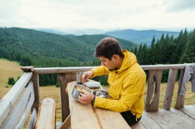Erkek turist, dağlardaki bir bungalovun balkonunda oturuyor ve arka planda makarna yiyor, ocakta soğukta yemek pişiriyor.