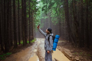 Sırtında sırt çantasıyla gezen genç bir turist, su birikintileriyle dolu bir yolda ıslak bir yağmur ormanında duruyor ve sisli bir zeminde yükseltilmiş roka ile akıllı bir telefondan bir ağ arıyor..