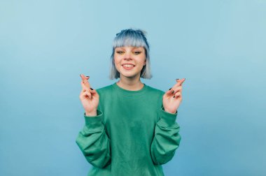 Mavi arka planda parmaklarını çapraz yapmış pozitif hipster kız kameraya bakar ve gülümser, bir dilek tutar.