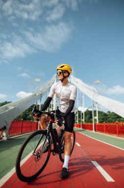 Profesyonel bir bisikletçi güneşli bir günde köprüde duruyor ve kameraya ciddi bir yüzle poz veriyor..