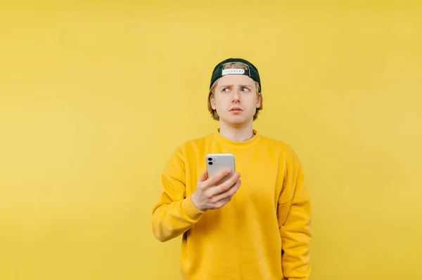 Young man in a yellow sweatshirt stands with a smartphone in his hands on a yellow background and looks away with a serious face