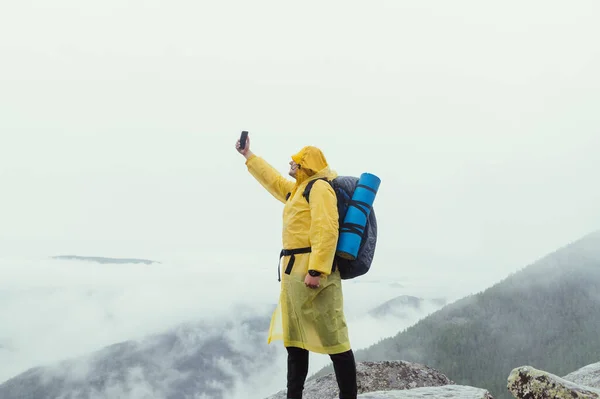 비옷을 배낭을 등산가는 날씨에 안개가 전망을 배경으로 산꼭대기에 스마트폰으로 비디오를 — 스톡 사진