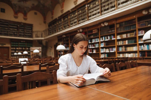 迷人的女人穿着白衬衫坐在图书馆的桌子旁 认真地读着一本书 — 图库照片