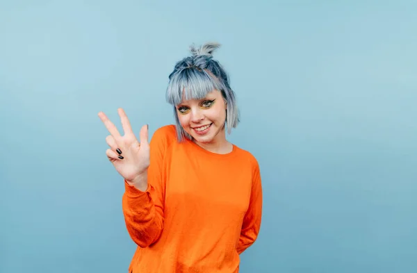 オレンジのセーターを着て 彼女の顔に笑顔で平和のジェスチャーを示す青の背景に立って青い髪を持つ正の女性 — ストック写真