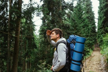 Sırtında sırt çantasıyla yürüyüş yapan bir erkek yürüyüşçü yüzünde bir gülümsemeyle ormanın ve patikaların arka planında.