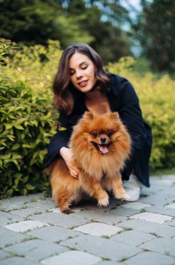 Yüzünde bir gülümseme olan pozitif bir kadın. Parktaki kaldırımda oturan sevimli turuncu bir köpekle..
