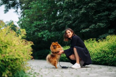 Mutlu esmer kadın parkta köpek cins Pomeranian Spitz ile oturuyor, yüzünde bir gülümsemeyle kameraya bakıyor..