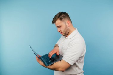 Tüylü yakışıklı yetişkin adam mavi arka planda dizüstü bilgisayardan hoşlanıyor ciddi bir yüz ifadesi ve beyaz tişört giyiyor. İzole edilmiş.