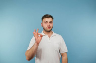 Beyaz tişörtlü, kıllı, yetişkin bir adam kameraya jest yapıyor ve dikkatlice mavi arka planda izole görünüyor.