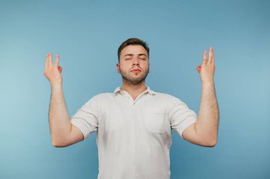Beyaz tişörtlü yetişkin adam, gözleri kapalı, kolları havada, mavi arka planda izole bir şekilde meditasyon yapıyor.