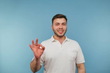 Tişörtlü neşeli adam mavi arka planda izole edilmiş, yüzünde gülümsemeyle kameraya iyi bir hareket gösteriyor..