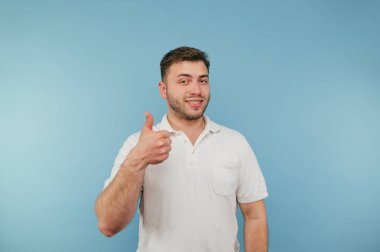 Beyaz tişörtlü olumlu genç adam mavi arka planda duruyor ve yüzünde bir gülümsemeyle bir jest gösteriyor..