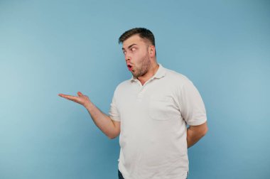 Beyaz tişörtlü genç adam mavi arka planda duruyor ve şok olmuş bir yüzle elini yana gösteriyor..